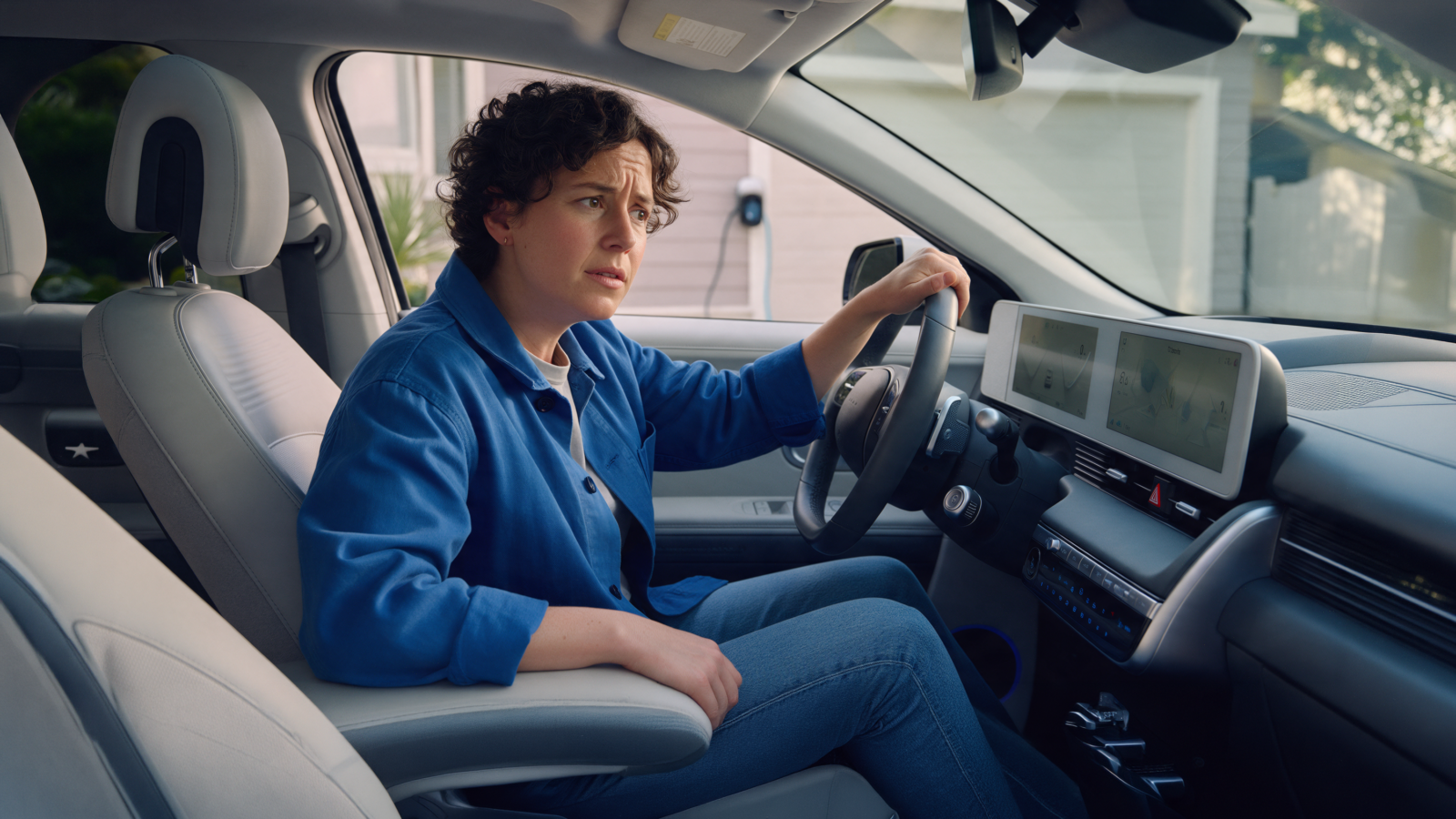 Woman sitting in the driver’s seat of an electric car with a worried expression, with a home EV charger visible outside.